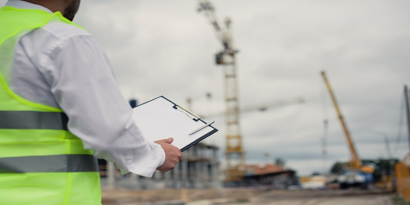 Chief in a vest and a construction helmet with documents in his hands, against the background of a tower crane and the sky with clouds. House construction inspection.